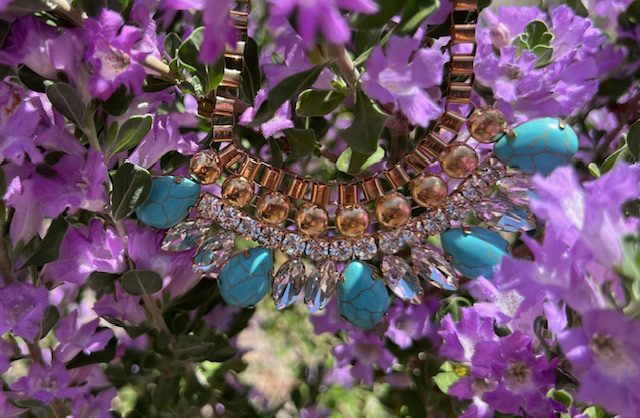 Ornate necklace with turquoise and clear stones against purple flowers.